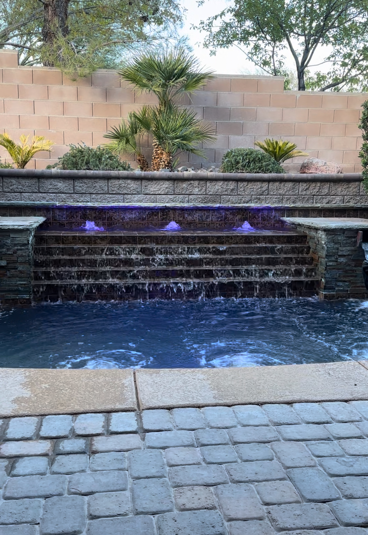 Pool waterfall feature with purple LED lighting and a landscaped backyard at dusk